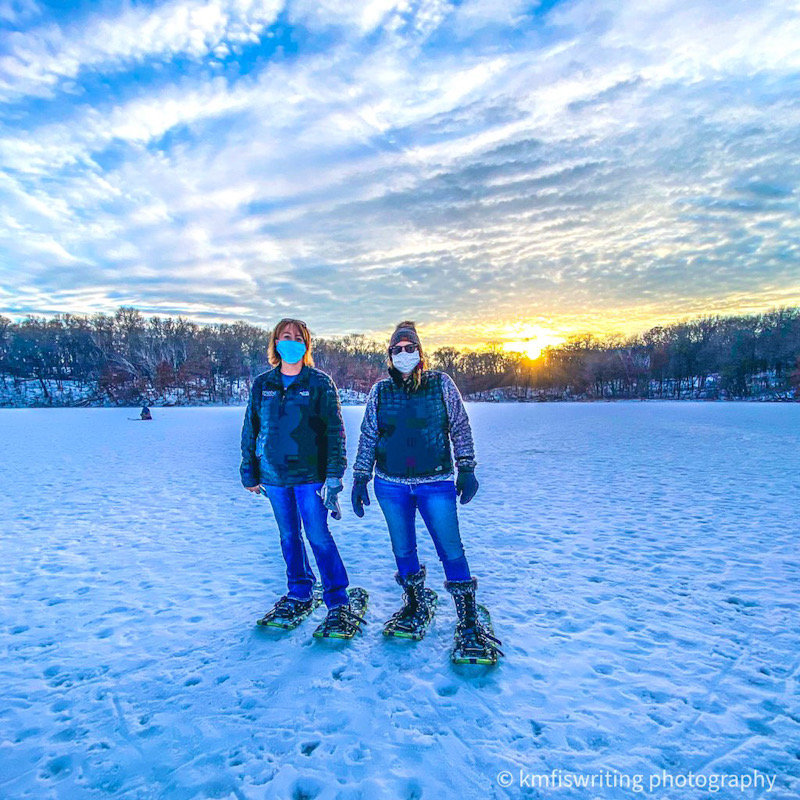 Two women wearing face masks while snoweshoeing on a frozen lake with a sunset in the background